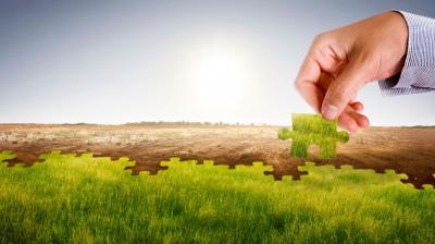 A hand places a green puzzle piece into a barren landscape, transforming it into grassland under a sunny sky.
