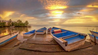 Three boats rest on a shore at sunset, with a vibrant orange and blue sky reflecting on the calm water, creating a tranquil and serene atmosphere.