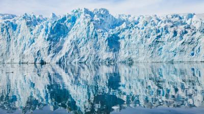 A glacier with jagged blue and white ice is reflected in calm, clear water under a partly cloudy sky.